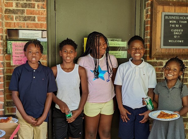 A group of 5 elementary aged youth are standing out in front of the LaFayette offices smiling and one has a paper plate with slice of pizza on it.